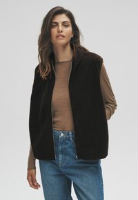 Brun fleeceväst med dragkedja fram, hög krage och inga ärmar, lagd över en ljusbrun topp, ihop med blå högmidjade jeans.