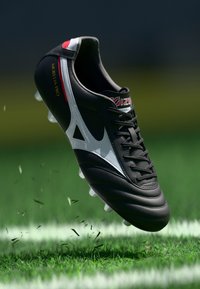 Melna futbola zābaki ar baltām detaļām, ādas virsma, paceltas šņores un seši caurspīdīgi tapu, kas atrodas virs zaļas zāles ar izkliedētu atkritumu.