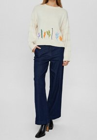 Maglione crema con ricami floreali multicolori all'orlo, abbinato a pantaloni a gamba larga blu scuro e stivaletti neri con tacco.
