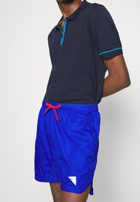 Polo shirt blu navy con colletto zip e accenti azzurri, abbinato a pantaloni da nuoto blu brillante con coulisse arancione e logo.