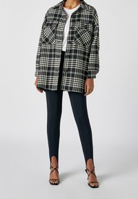 Chemise surdimensionnée à carreaux noirs et blancs avec deux poches poitrine, associée à des leggings noirs et des talons noirs à brides.