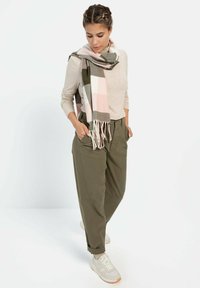 Lichtbeige lange mouw top, olijfgroene broek en een roze, groene en witte geblokte sjaal met franjes. Beige sneakers maken de outfit compleet.