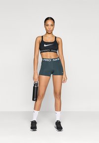 Czarny stanik sportowy z białym logo Nike, zielone spodenki z gumką w napisy "PRO NIKE", czarna butelka na wodę, białe skarpetki oraz czarne buty sportowe.