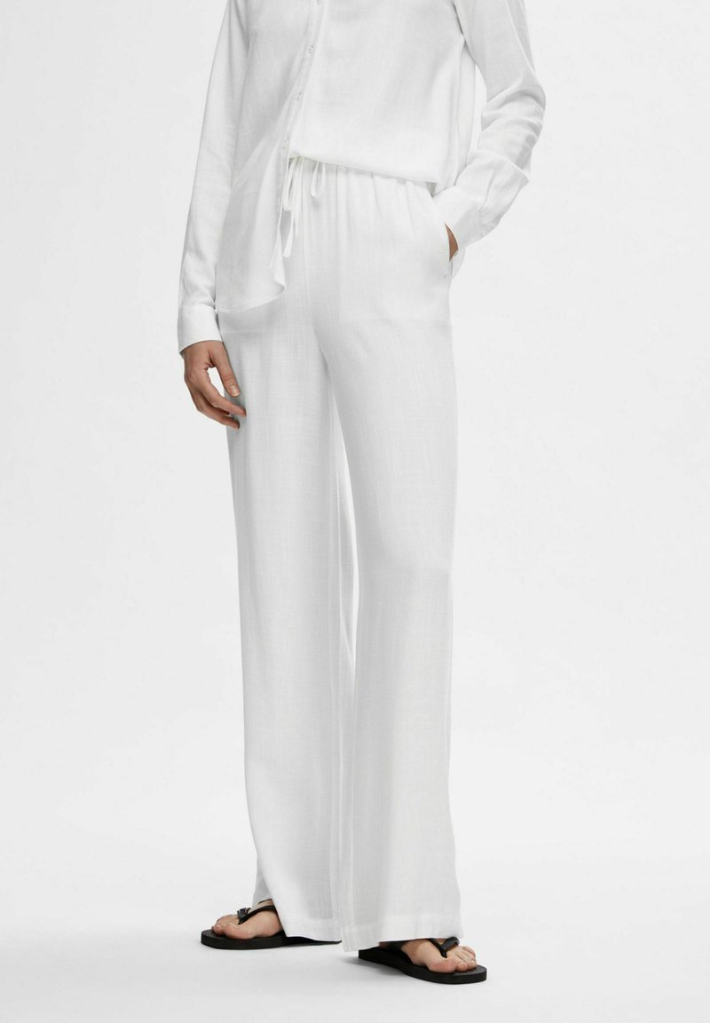 Witte linnen broek met een losse pasvorm en een elastische tailleband, gecombineerd met een bijpassend langhemd. Flat zwarte sandalen maken de outfit compleet.