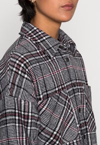 Close-up van een persoon die een zwart, wit en rood geruite blouse met kraag en een voorzak draagt, waarbij de lippen en kaaklijn zichtbaar zijn.