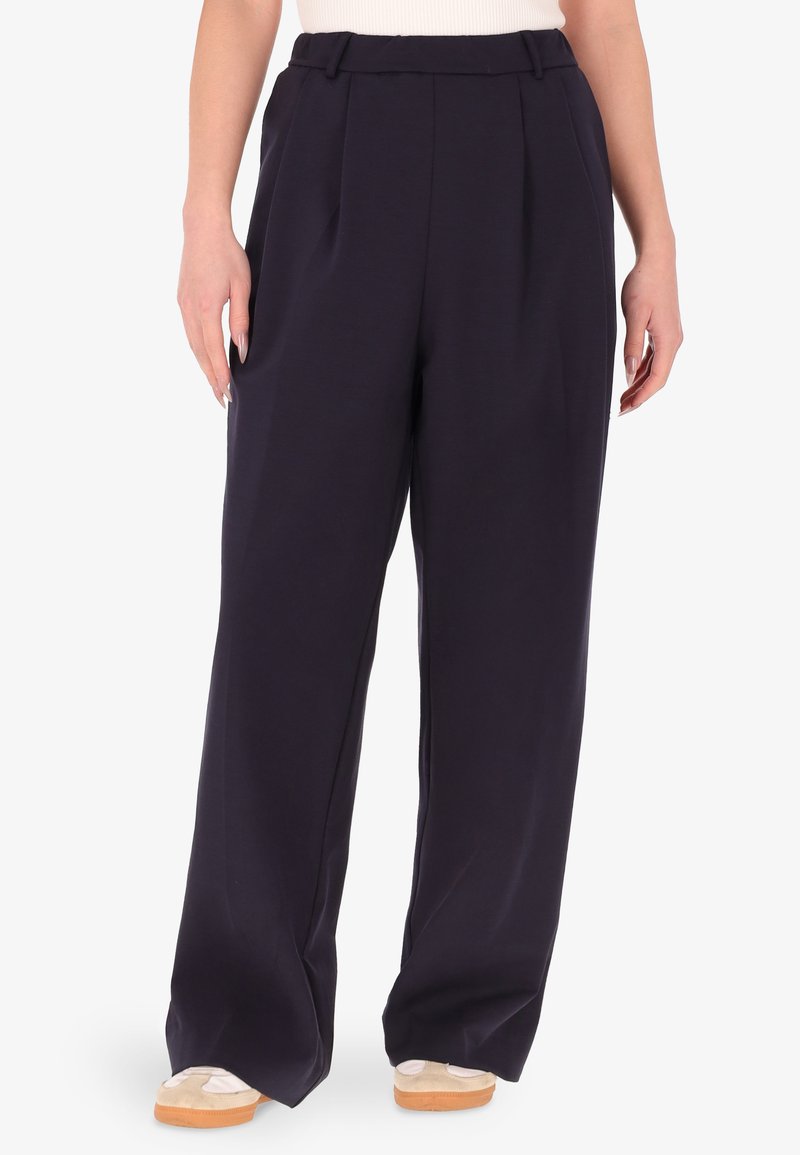 Pantaloni larghi di un blu scuro con una parte frontale pleat, vita elasticizzata e una consistenza del tessuto morbida, abbinati a scarpe slip-on beige.