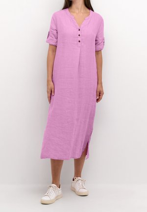 Vestido rosa de lino con cuello en V, mangas remangadas y botones en la parte delantera. Cuenta con una abertura lateral y llega hasta la mitad de la pantorrilla. Se lleva con zapatillas blancas.