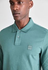 Polo verde confecționat dintr-un material texturat, cu guler, două butoane și un mic logo brodat pe pieptul stâng.