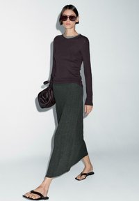Langer dunkelgrüner Ripprock, kombiniert mit einem eng anliegenden dunklen Oberteil und schwarzen Sandalen. Das Model trägt eine dunkelbraune Tasche. Große rechteckige Sonnenbrille auf. 