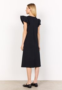 Frau steht seitlich in einem schwarzen Midikleid mit fransigen Ärmeln und schwarzen Mary Jane Schuhen vor einem schlichten weißen Hintergrund.