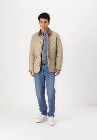 Tan quilted jacket with brown corduroy collar, button front, and side pockets. Worn over a light blue shirt and paired with blue jeans and white sneakers.