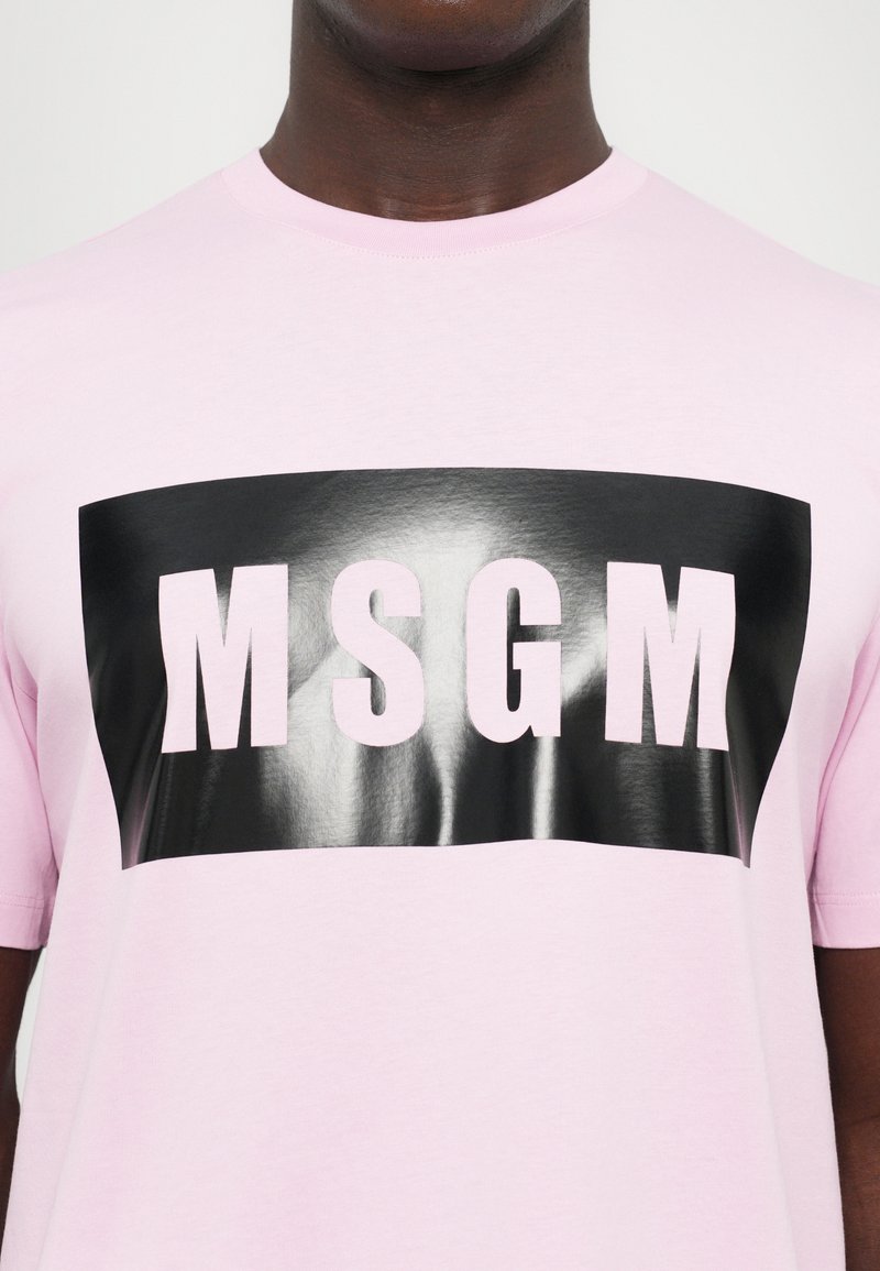 Man wearing light pink T-shirt with a glossy black rectangular patch displaying large, bold letters "MSGM" in light pink.