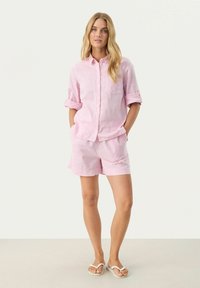 Lyserød linned skjorte med knapper og oprullede ærmer, parret med matchende lyserøde shorts. Model står på en lyse baggrund.