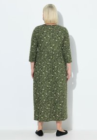 Abito maxi in tessuto verde scuro con stampa floreale, maniche a tre quarti e vita aderente con gonna fluida. Scarpe nere piatte.