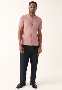 Camicia rosa a maniche corte con colletto e chiusura frontale con bottoni, abbinata a pantaloni neri larghi e scarpe nere. Design semplice e tessuto liscio.