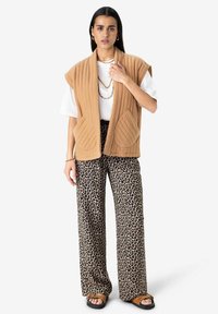 Beige quilted vest with wide collar and two front pockets, paired with leopard print wide-leg pants and sandal shoes. White t-shirt underneath.