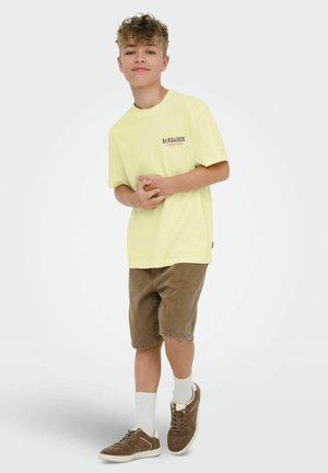Boy with curly hair wearing a yellow "Barbados" T-shirt, brown shorts, white socks, and brown sneakers, standing against a plain background.