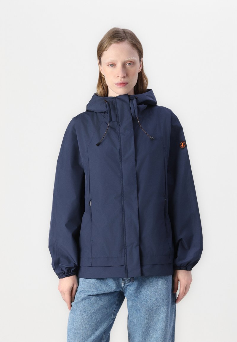 Giacca leggera blu navy con zip frontale, cappuccio regolabile e polsini elasticizzati. Presenta tasche laterali e un badge con logo sulla manica.