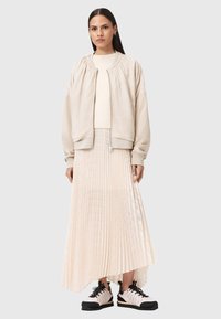 Giacca bomber beige con collo arricciato, top chiaro e gonna plissettata fluida a lunghezza variabile in tonalità morbide, abbinata a sneaker nere e rosa.