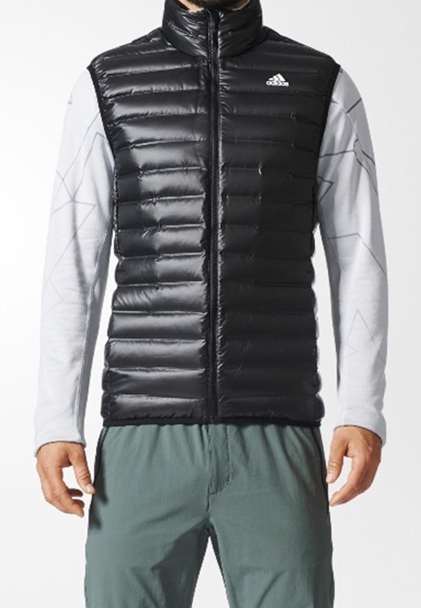 Mann trägt eine schwarze gepolsterte ärmellose Weste mit Adidas-Logo über einem hellgrauen Langarmshirt und grünen Hosen.