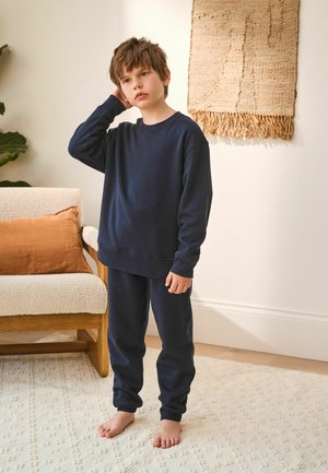 Navy blue sweatshirt and matching sweatpants, cotton material, relaxed fit, ribbed cuffs, worn barefoot, standing in a cozy room.