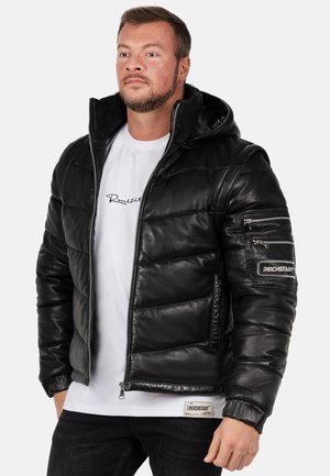 Man draagt een zwarte gewatteerde leren jas over een wit T-shirt met print, staat met handen langs het lichaam, kijkt vooruit tegen een witte achtergrond.