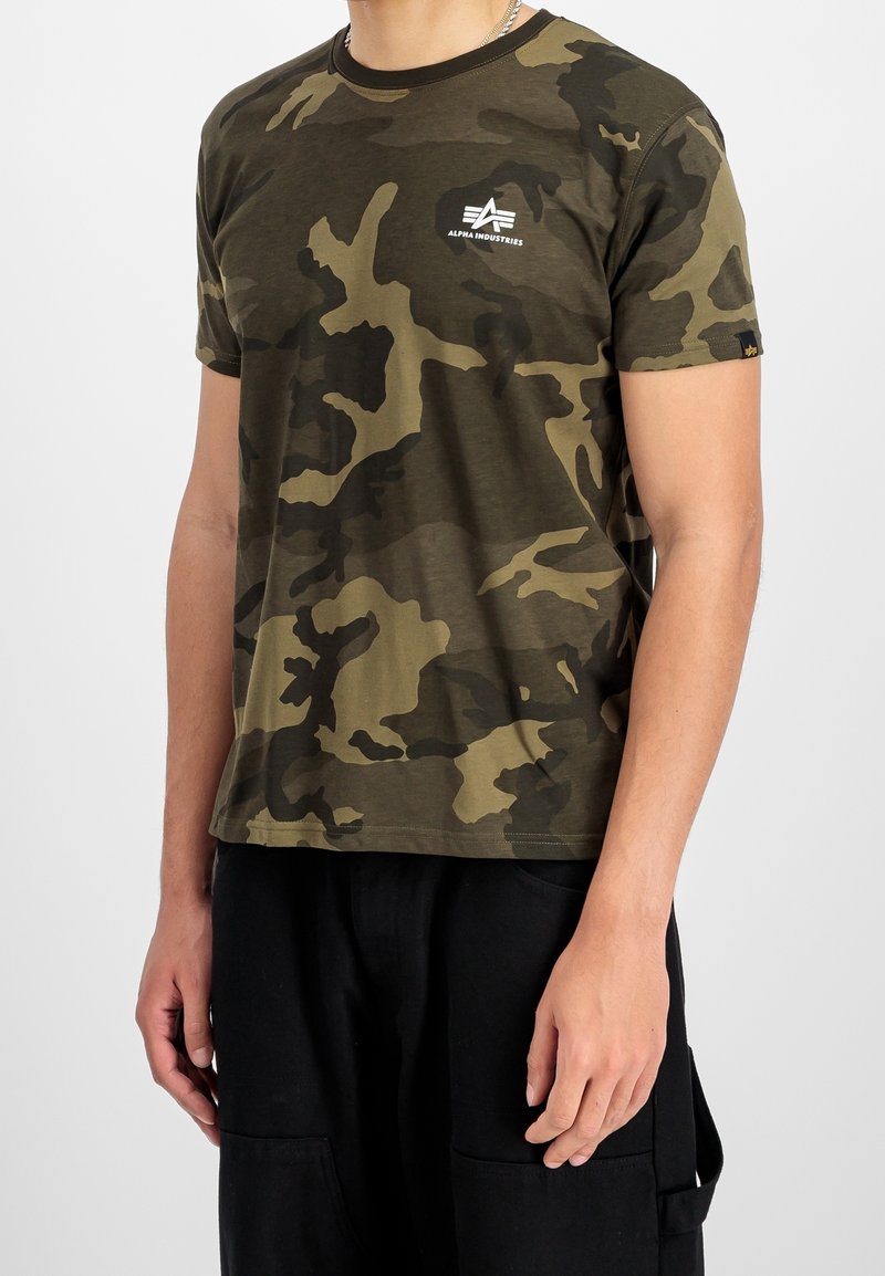 Homme portant une chemise à manches courtes camouflage verte et marron de la marque Alpha Industries avec un pantalon noir sur un fond uni.