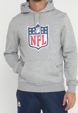 Mężczyzna w szarej bluzie z kapturem z logo NFL i kieszenią z przodu oraz granatowych spodniach Kappa na jednolitym białym tle.