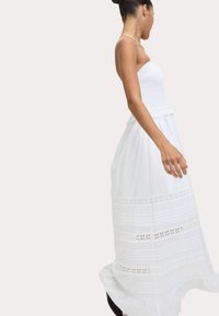 Abito maxi bianco con scollo arricciato, gonna svasata e bande orizzontali ricamate. Tessuto leggero e fluido con leggero volants all'orlo.