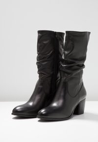 Botas de tobillo de cuero negro con un diseño holgado, cremallera lateral, punta redondeada y un tacón cuadrado de altura media. Textura suave con acentos mínimos.