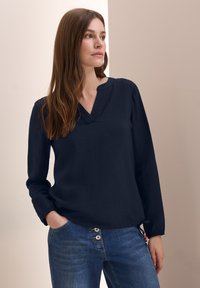 Marineblauwe blouse met lange mouwen, een V-hals en een gestructureerde stof. Gepaald met lichtblauwe jeans met een knoopdetail in de taille.