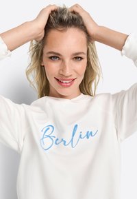 Bijela dukserica s velikim plavim izvezenim natpisom "Berlin". Mekana tkanina s opuštenim krojem i okruglim izrezom.