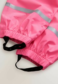 Lyserøde vandtætte bukser med elastiske manchetter og reflekterende sølvstriber, der har sorte justerbare fodstropper på en lys baggrund.