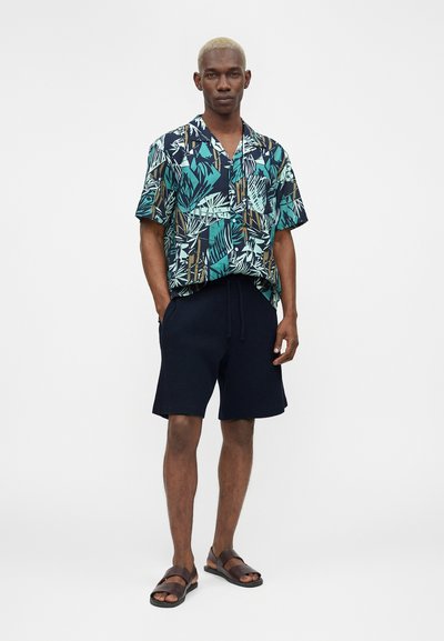 Άνδρας με κοντά ξανθά μαλλιά, φοράει μπλε, πράσινο και μπεζ πουκαμίσα με σχέδια και κοντό μανίκι, σκούρα μπλε σορτς και καφέ σανδάλια, στέκεται με τα χέρια στις τσέπες.