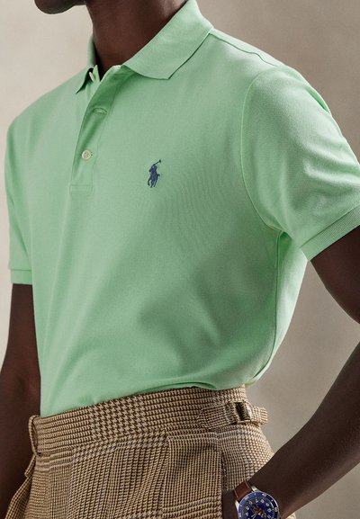 Ανοιχτό πράσινο πόλο πουκάμισο από υφασμάτινη υφή με γιακά τριών κουμπιών και μικρό ναυτικό λογότυπο. Φορεμένο με καρό παντελόνια και ρολόι.