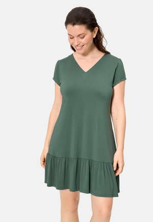 Mujer con vestido corto de manga corta, escote en V y dobladillo fruncido de color verde oscuro, mirando hacia abajo y sonriendo frente a un fondo claro y simple.