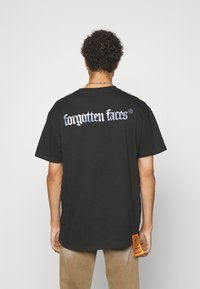 Svart oversized t-skjorte med teksten "glemte ansikter" i blå gotisk skrifttype på baksiden, og en oransje merkeetikett på siden. Myk tekstur.