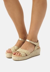 Sandales compensées avec un dessus en paille tressée, une sangle de cheville en métal doré et une plateforme en jute. Le design à bout ouvert met en valeur les pieds.