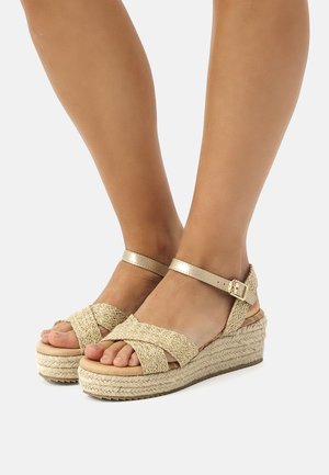 Sandalias de cuña con parte superior de paja trenzada, tirante metálico dorado en el tobillo y plataforma envuelta en yute. El diseño de punta abierta destaca los pies.