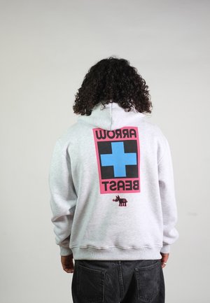 Persona con capelli ricci che indossa una felpa con cappuccio grigio chiaro con una grande croce blu e testo "ARROW BEAST" rovesciato sulla schiena.