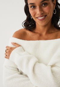 Off-the-shoulder hvid striktrøje med ribbet struktur og lange ærmer. Modellen holder stoffet, som har en subtil glans, og viser det bløde materiale.