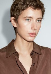 Camicia in satin marrone con colletto, caratterizzata da un leggero lucido e bottoni. La camicia è indossata da una persona con capelli corti e occhi azzurri.