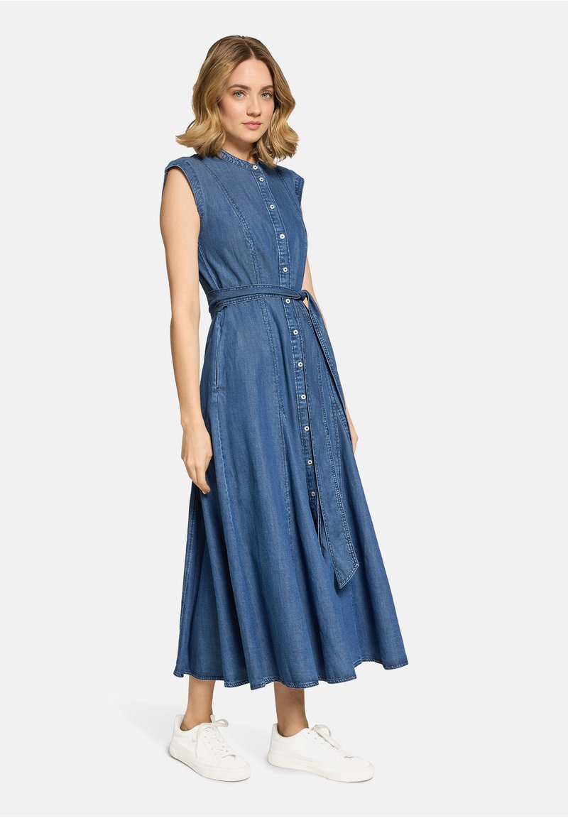 Femme portant une robe sans manches en jean bleu avec boutons et ceinture à la taille, ainsi que des baskets blanches, debout devant un fond clair.
