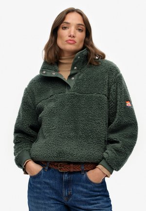 Groene fleece pullover met hoge kraag, drukknoopsluiting en een borstzak. Kenmerkt zich door een gestructureerde stof en wordt gecombineerd met blauwe jeans en een bruine riem.