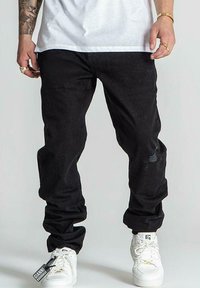 Pantalones de mezclilla negros con un corte relajado, con ligeras desgastaciones. Combinados con zapatillas blancas, mostrando una etiqueta adjunta a los pantalones.
