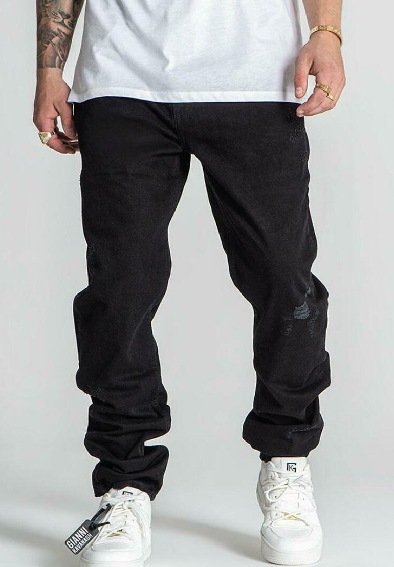 Pantalones de mezclilla negros con un corte relajado, con ligeras desgastaciones. Combinados con zapatillas blancas, mostrando una etiqueta adjunta a los pantalones.