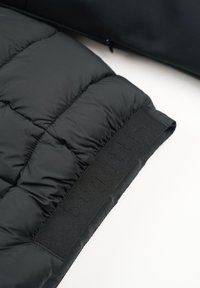 Schwarze Daunenjacke aus Stoff mit gestepptem Muster. Verfügt über einen mattschwarzen Klettverschluss mit geprägtem Branding. Weiches und isolierendes Material.