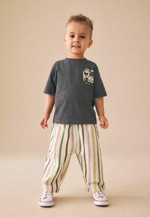 Väikelaps seisab, seljas tumehall särk, millel on veoauto pilt, ja triibulised lahtised püksid, naeratab ühtlase beeži tausta ees.