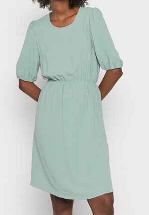 Woman wearing a light green, knee-length dress with puff sleeves and an elastic waistband, standing with hands behind her back.