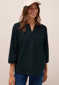 Blusa in cotone verde scuro con scollatura a V e maniche 3/4. Presenta polsini arricciati e una vestibilità morbida, abbinata a jeans blu.
